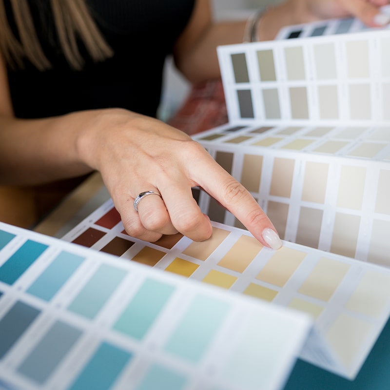 Main d'une personne pointant une nuance beige sur un nuancier de couleurs murales. Autres palettes visibles pour la décoration intérieure.