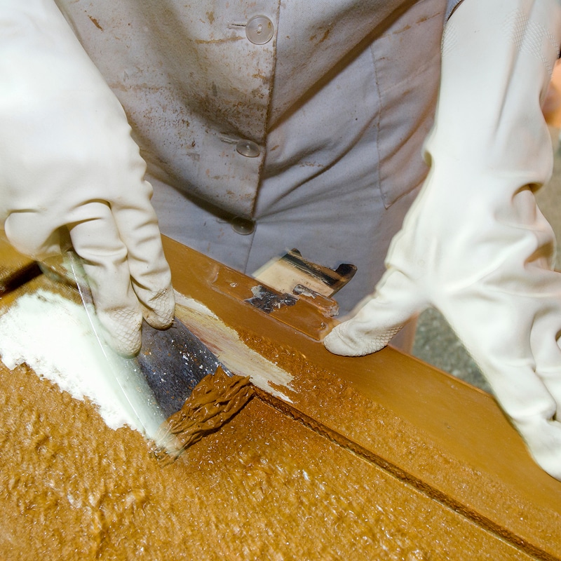Personne gantée décapant une épaisse couche de peinture brune d'une surface en bois. Un produit blanc et un racloir sont utilisés, révélant le bois foncé.