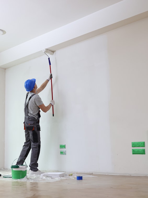 Ouvrier peintre en salopette et casque bleu utilise un rouleau à manche pour peindre un mur blanc. Matériel de peinture au sol.