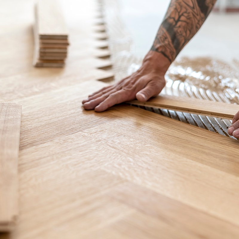 Main d'un artisan posant des lattes de parquet en bois clair motif chevrons sur un sol encollé. Stacks de lames en arrière-plan.