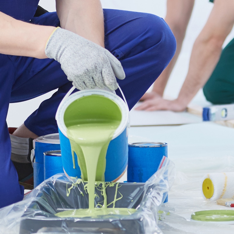 Main gantée d'un artisan en bleu versant de la peinture vert clair d'un pot dans un bac à rouleau, préparant le chantier.