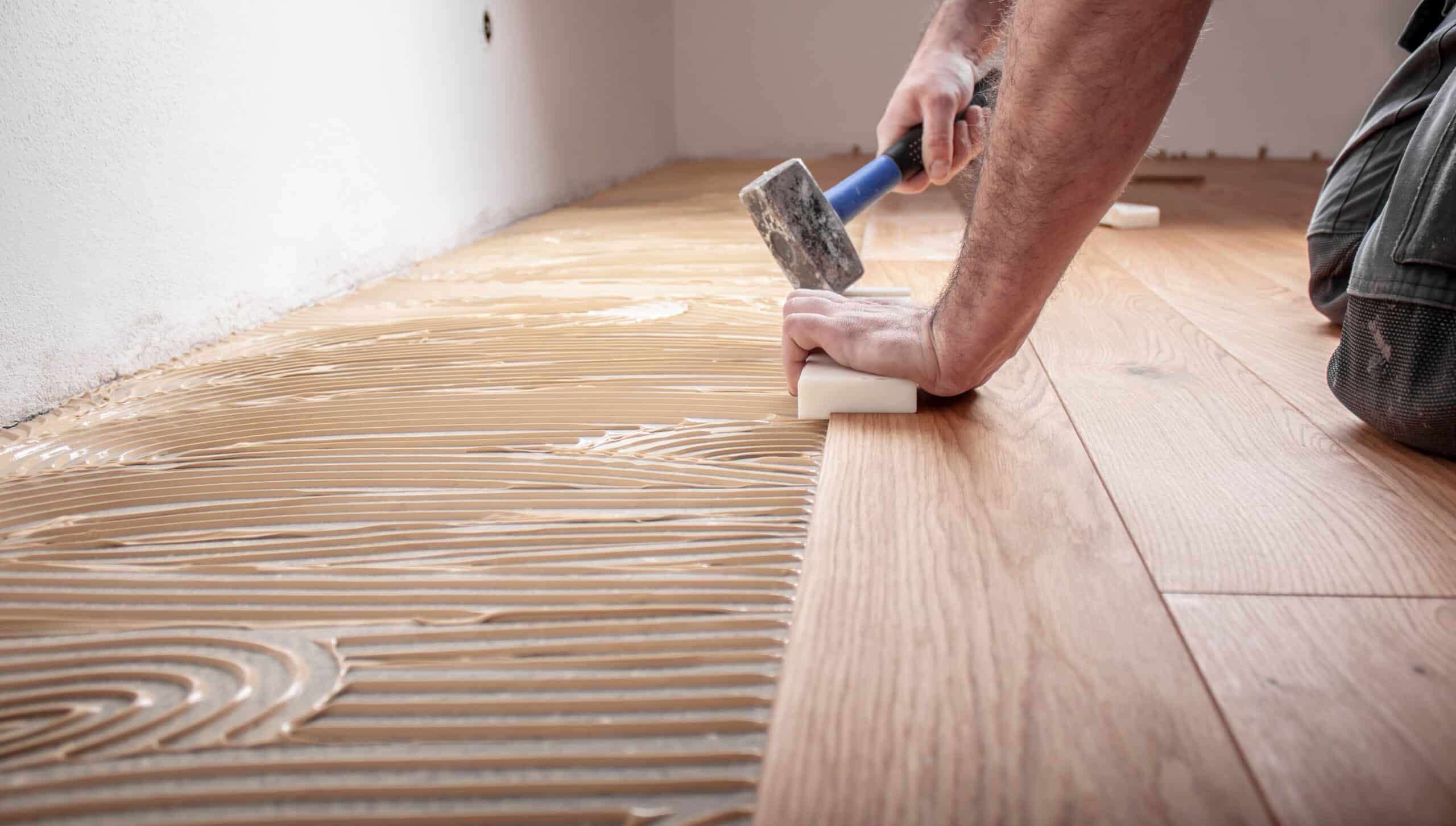 Homme posant du parquet en bois sur un sol enduit de colle. Il utilise un maillet et un bloc de frappe pour ajuster les lames de bois.