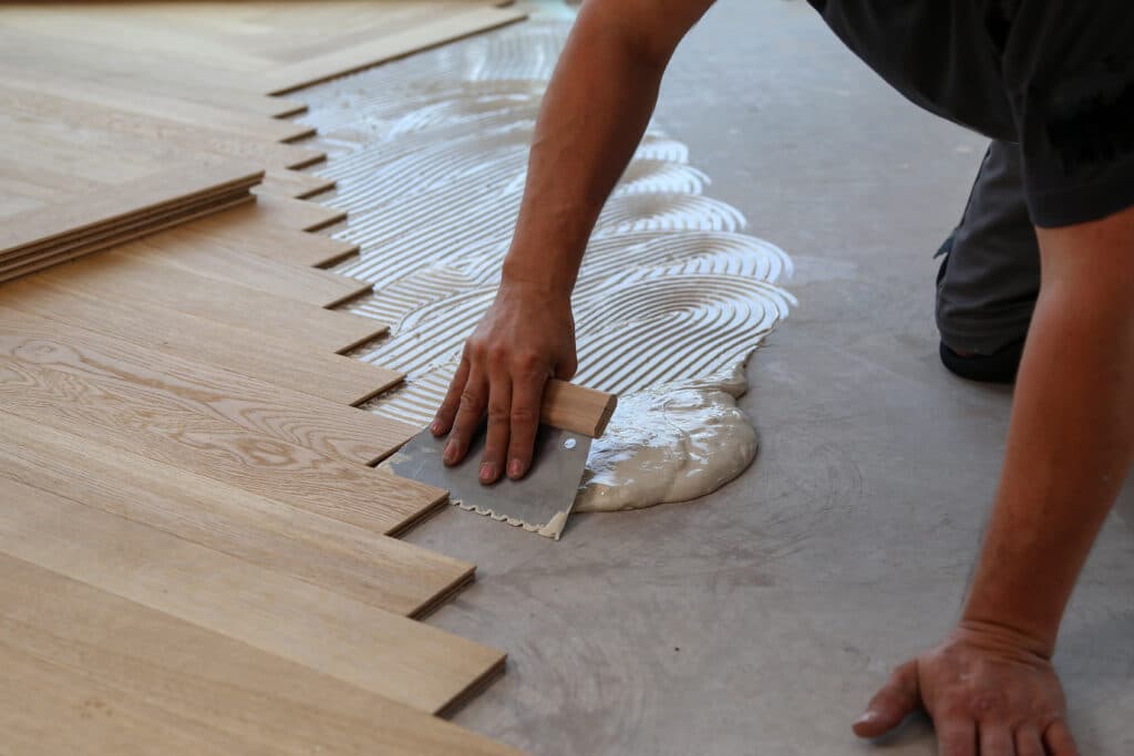 Pose de Parquet en Bois: Étapes d'Application de la Colle Ouvrier étalant de la colle sur un sol en béton avec une spatule crantée pour la pose de parquet en bois clair. Lames de parquet prêtes.