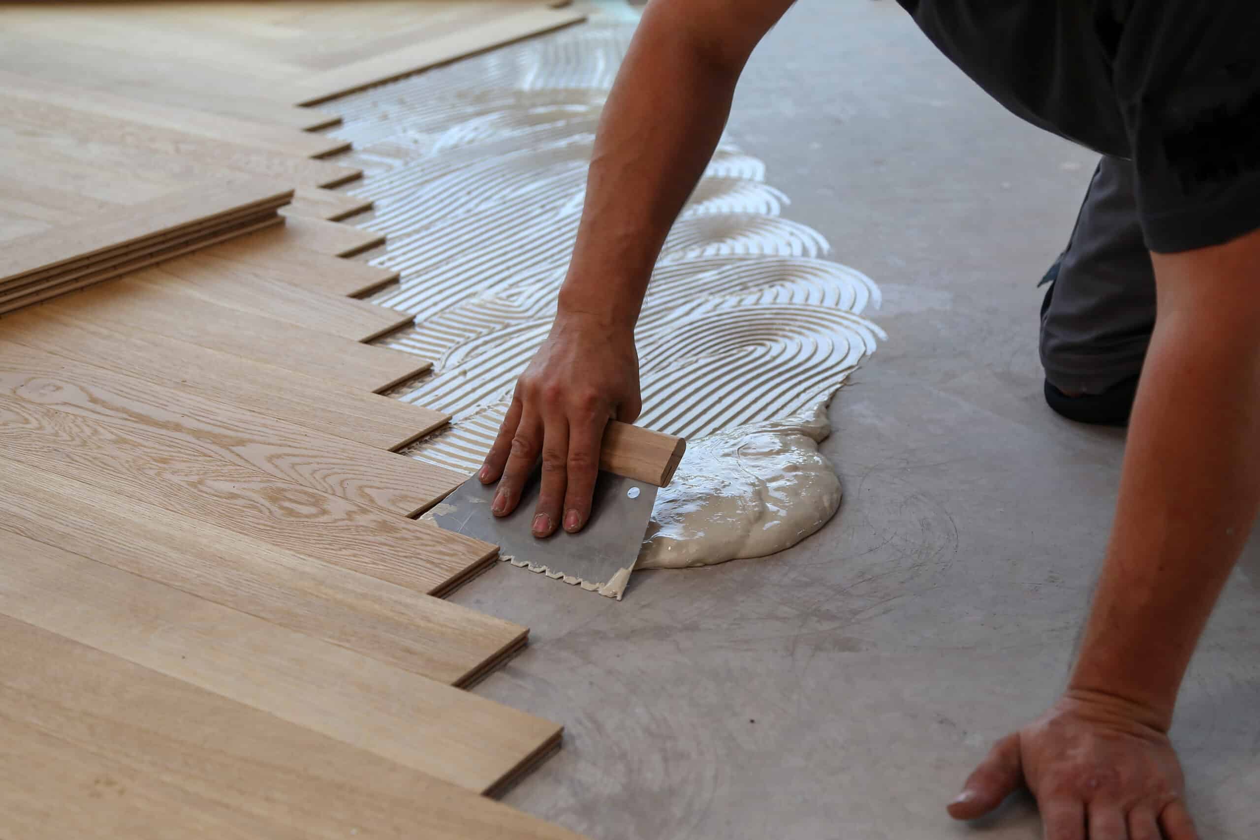 Ouvrier étalant de la colle sur un sol en béton avec une spatule crantée pour la pose de parquet en bois clair. Lames de parquet prêtes.